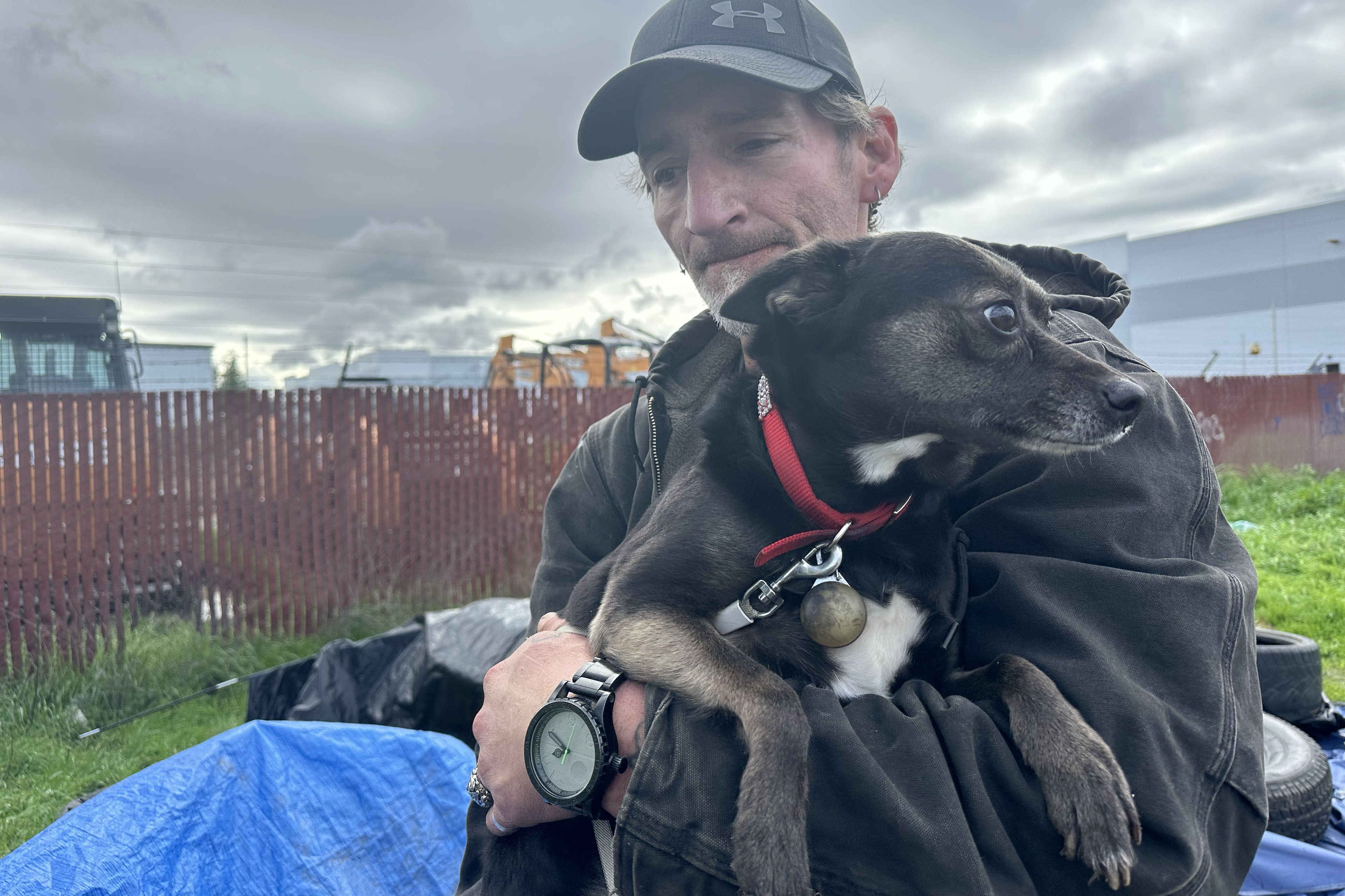 Samuel Buckles stands outside and holds his dog in his arms. The dog is a small mixed-breed with black fur and a few white spots on its underside.